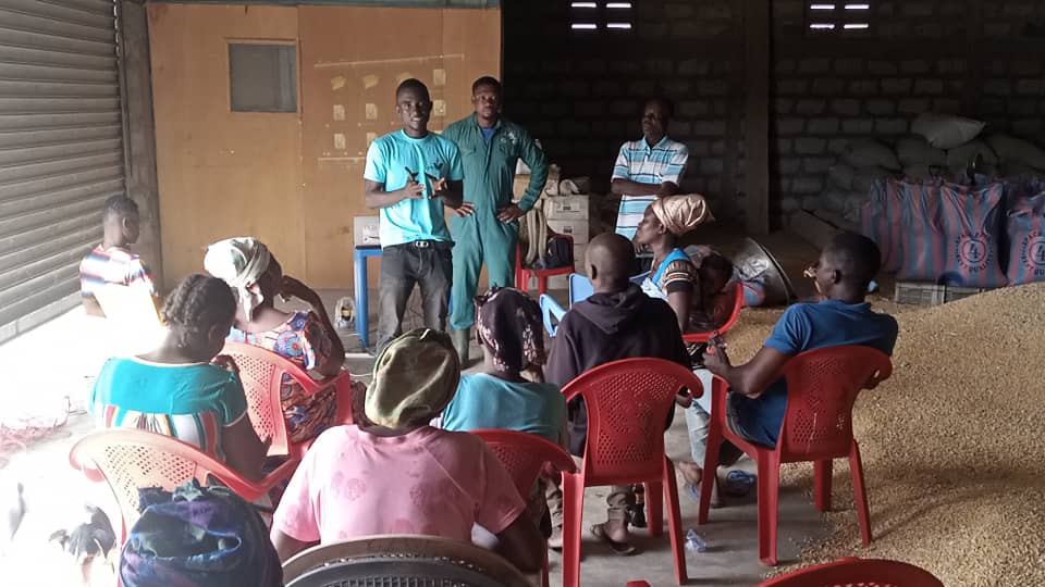 Farmers seated on red plastic chairs during a session in a storage building, with facilitators standing at the front, image 8 of 10