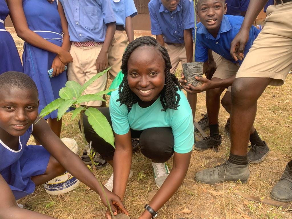 A smiling facilitator in a teal T-shirt planting a seedling surrounded by pupils in school uniform, image 1 of 14
