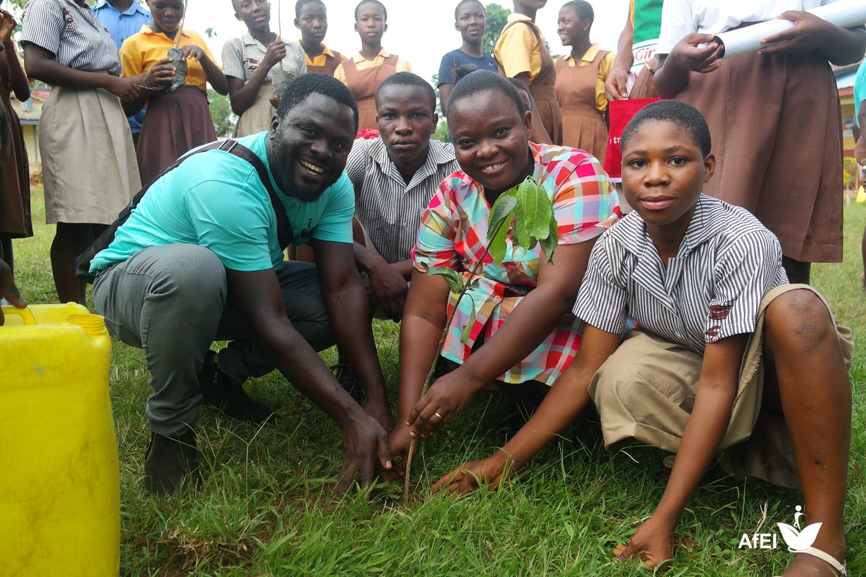 Pupils, students and facilitators in teal T-shirts kneeling together to plant a tree seedling on a school field