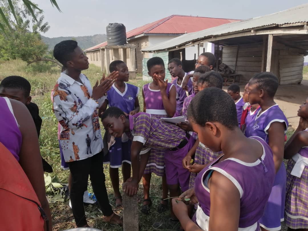 Pupils in purple school uniform gathered around a facilitator in a patterned shirt outdoors, image 14 of 14