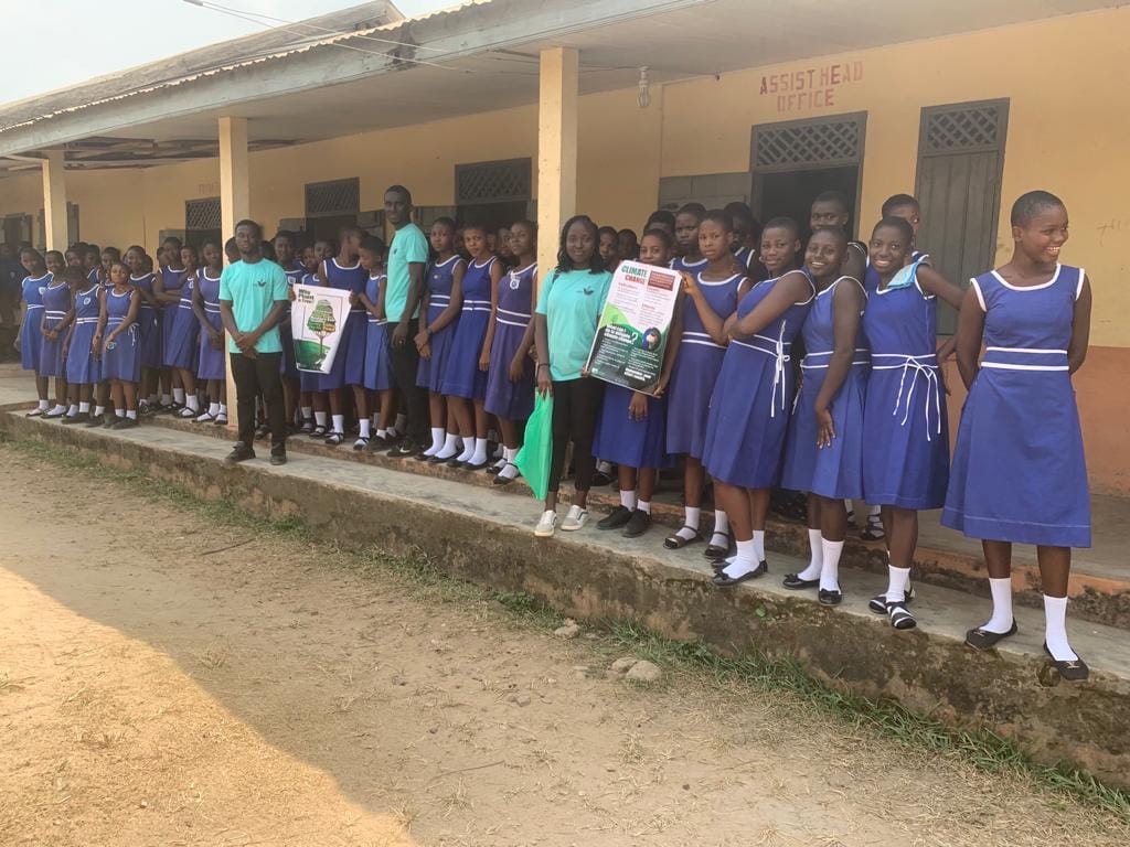 Pupils in blue school uniform standing in a line outside a school building, some holding tree-planting posters, image 2 of 14