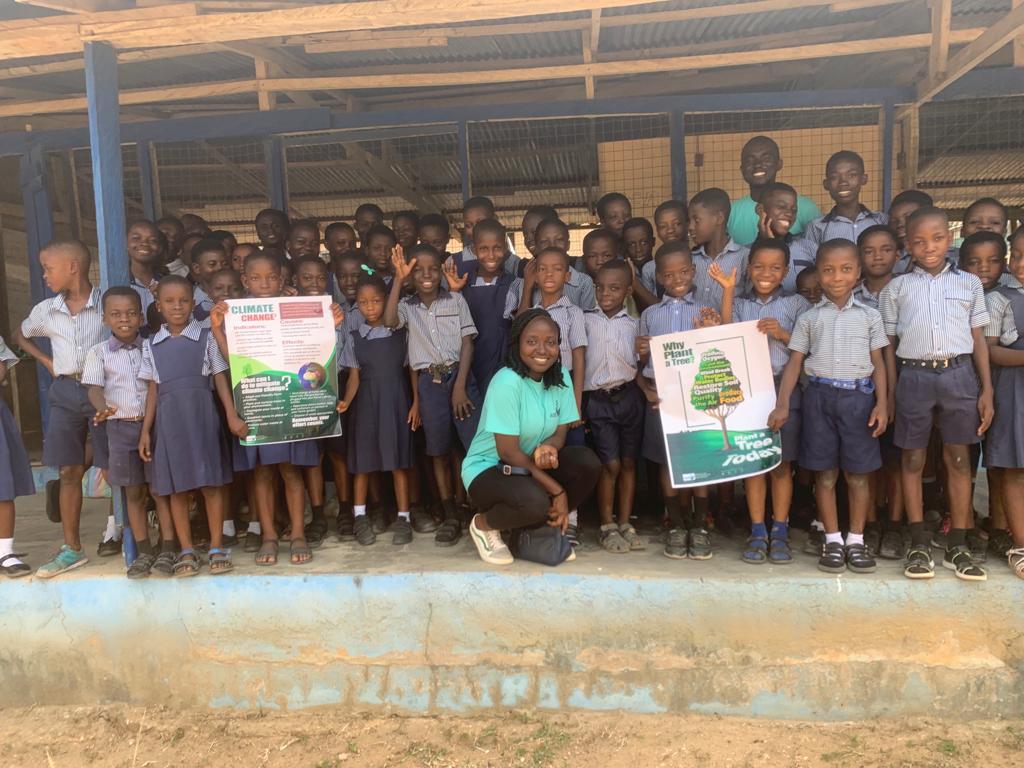 Young pupils in school uniform gathered in rows on a school veranda, two people holding tree-planting posters, image 6 of 14